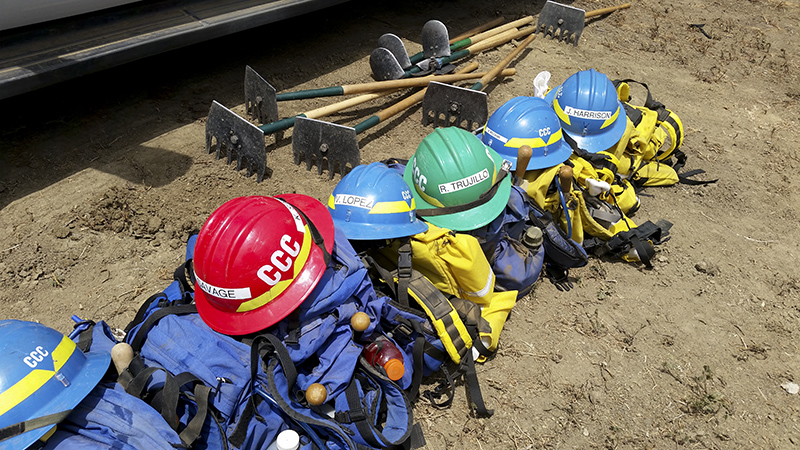 Helmets-and-tools-Fire-crew-FEATURED-IMAGE Corpsmembers with the Placer 1 fire crew cut fire line with hand tools as flames from the Dixie Fire approach in 2021.