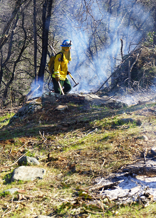 SideImage_Magalia_500x700 Corpsmember next to burn pile, with hand tool.