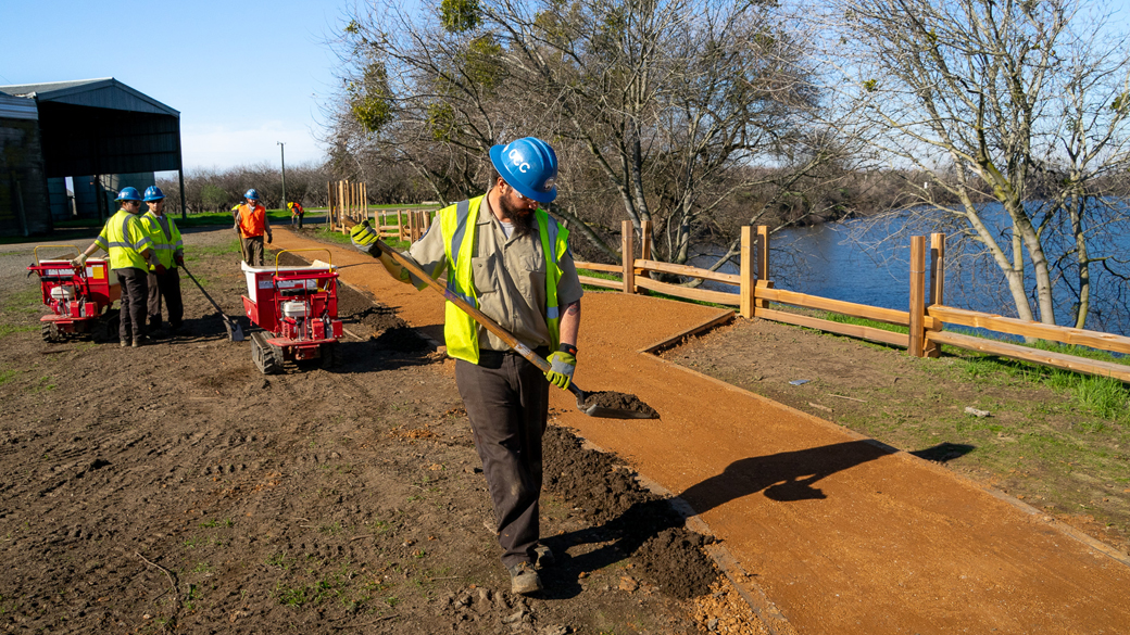 male holding shovel with dirt sprinkling it on edge of trail with other people off in the distance.