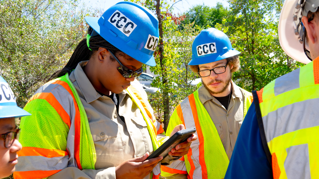 female wearing helmet looking at tablet, with male watching her work