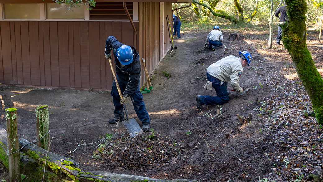 corpsmembers use hand tools around perimeter of a building clearing every bit of vegetation