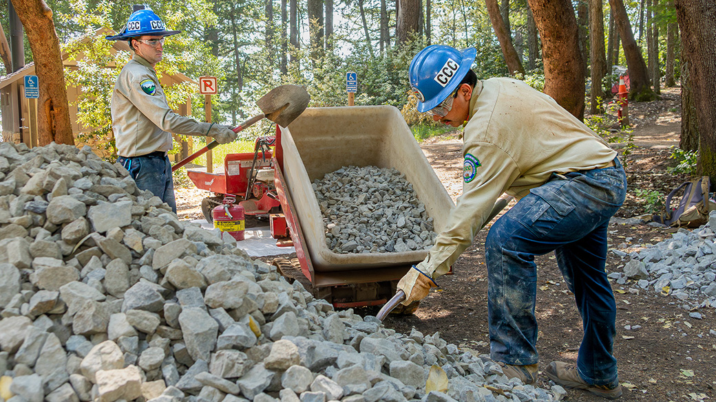 two male corpsmembers with shovels moving rock from a pile to a motorized wheelbarrow