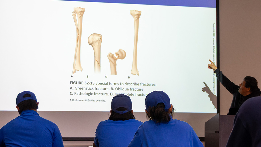 lecturer pointing at screen as three corpsmembers look on, screen shows different broken limbs in illustrations