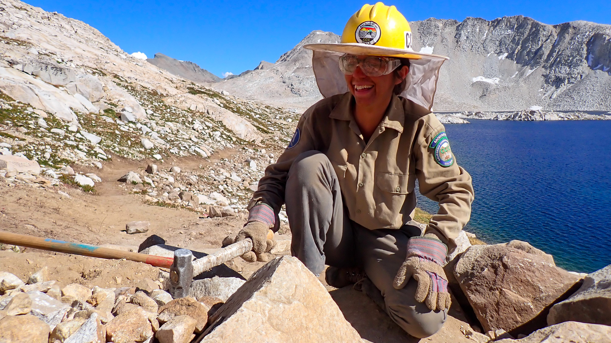 female smiles while holding a hammer in front of an alpine lake