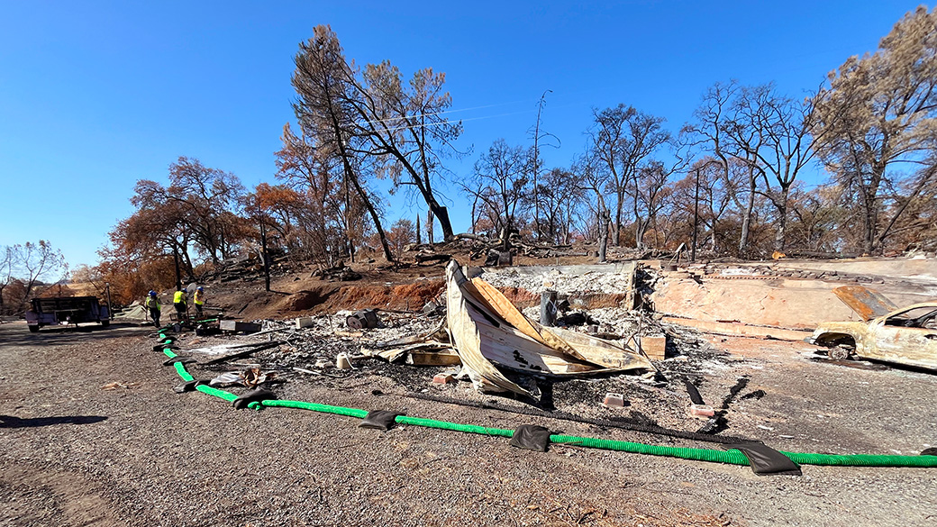 green silt sock surround a burned home, with corpsmembers working in the far distance