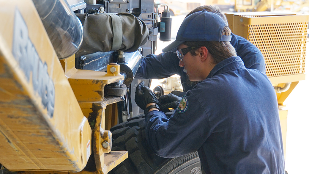 male in blue hat and coveralls working on a large mower in a garage